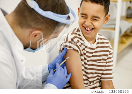 Male doctor is doing a vaccination. Man wearing white medical gown. Boy wearing brown plaid t-shirt. 133668196