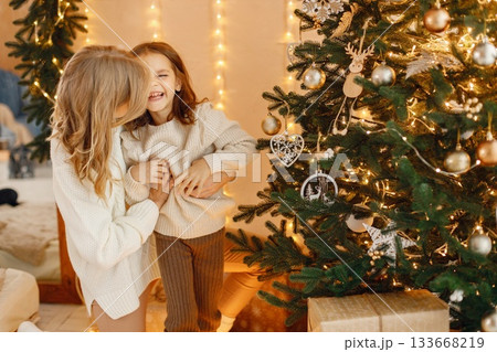 Portrait of a little girl celebrating Christmas with her mom at home. Girl and her mother having fun while standing near Christmas tree. Family wearing beige knitted clothes. Portrait of a little girl celebrating Christmas with her mom at home. Girl and her mother having fun while standing near Christmas tree. Family wearing beige knitted clothes. 133668219