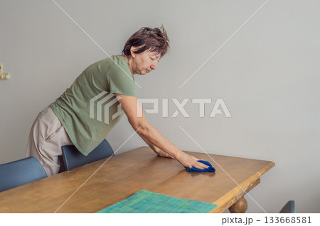 Unaltered senior woman cleaning and organizing a table at home, creating a tidy and comfortable living space. Concept of everyday routine, home care, cleanliness, wellbeing, responsibility and simple 133668581