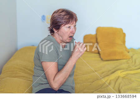 Unaltered senior woman brushing her teeth while sitting on her bed during her calm morning routine. Concept of self-care, hygiene, healthy aging, everyday wellbeing, slow living and authentic home 133668594