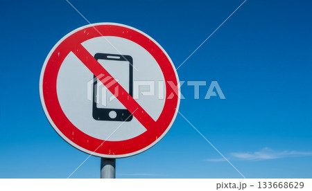 Round prohibition sign with a smartphone icon crossed out by a red diagonal line, indicating no mobile phones allowed or signal issues, set against a clear blue sky 133668629