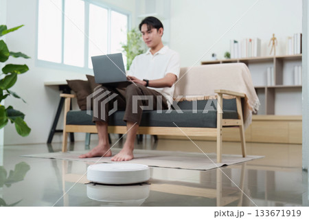 Remote Work. User using laptop on couch while robotic vacuum cleans nearby. Remote Work. User using laptop on couch while robotic vacuum cleans nearby. 133671919