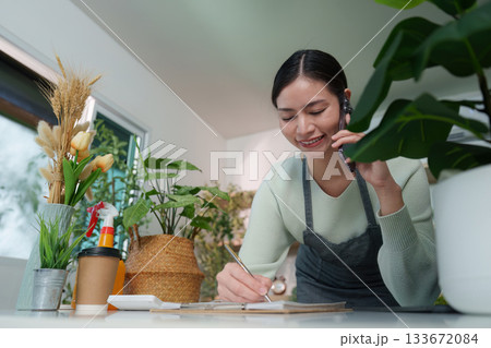 Business Call. Woman writing notes while talking on the phone in plant-filled space. 133672084