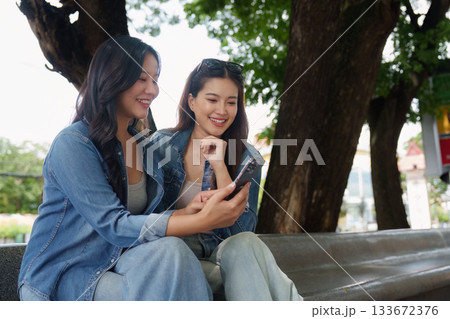 Casual Friendship. Two young women sitting together on a bench, sharing a smartphone and enjoying coffee in a park. 133672376