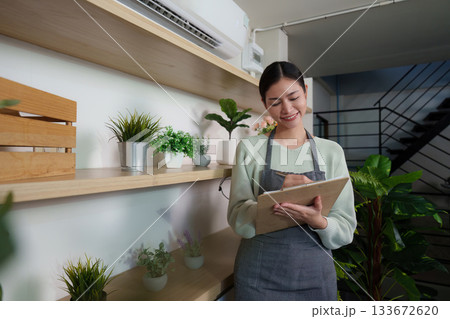 Sustainable Gardening. Smiling woman taking notes in a plant shop, promoting eco-friendly practices. Sustainable Gardening. Smiling woman taking notes in a plant shop, promoting eco-friendly practices. 133672620