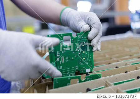 Worker in protective gloves placing green printed circuit board into cardboard tray for safe storage and shipping in electronics plant 133672737