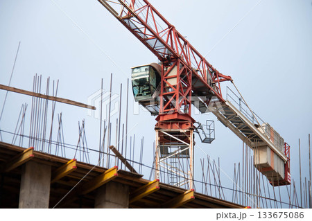 Close-up of Control Cabin of Red Tower Crane with Counterweight at Construction Site. In Foreground are Yellow Formwork Beams with Monolithic Box Fittings. 133675086