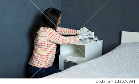 Young woman fixes an electrical outlet next to the bed at home working carefully with gloves on to ensure safety during the repair  133676841