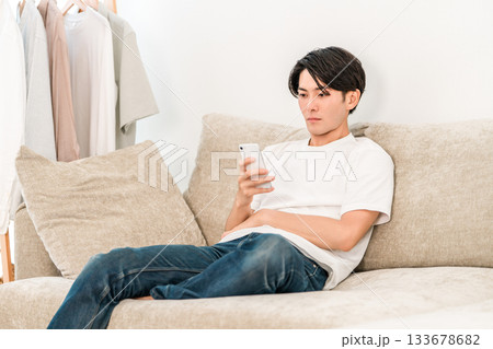 リビングソファーに座りスマホを見る若いアジア人男性（怠ける,ダラダラ） 133678682