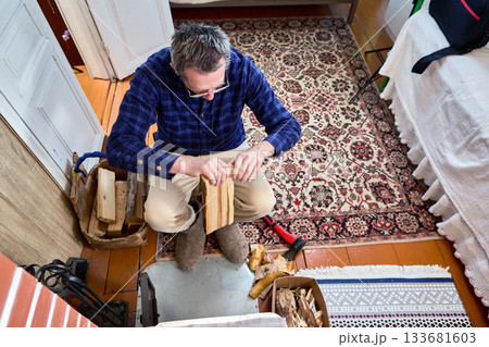 Splitting thin splinters from firewood piece, middle aged man prepares kindling supply for lighting brick hearth used for home heating. Splitting thin splinters from firewood piece, middle aged man prepares kindling supply for lighting brick hearth used for home heating. 133681603