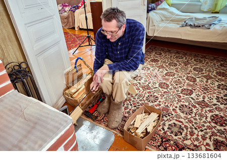 Mature man sits in front brick stove opening holding poker. 133681604