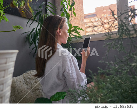 Woman in Robe Checking Phone in Cozy Balcony Garden Woman in Robe Checking Phone in Cozy Balcony Garden 133682639