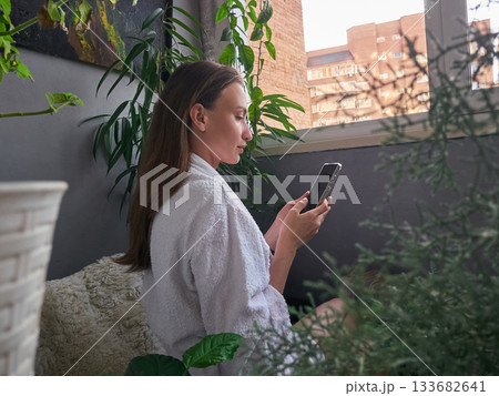 Woman in Robe Checking Phone in Cozy Balcony Garden 133682641