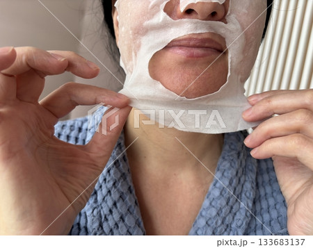 Mature woman applying a moisturizing sheet facial mask at home. Anti aging skincare routine for mature skin, hydration, rejuvenation, fine lines treatment, beauty self care concept 133683137