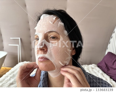 Mature woman applying a moisturizing sheet facial mask at home. Anti aging skincare routine for mature skin, hydration, rejuvenation, fine lines treatment, beauty self care concept Mature woman applying a moisturizing sheet facial mask at home. Anti aging skincare routine for mature skin, hydration, rejuvenation, fine lines treatment, beauty self care concept 133683139
