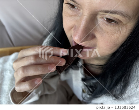 Woman sitting indoors biting a piece of dark chocolate, relaxed casual moment enjoying a sweet treat in a cozy home environment Woman sitting indoors biting a piece of dark chocolate, relaxed casual moment enjoying a sweet treat in a cozy home environment 133683624