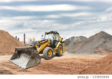A yellow backhoe loader moves dirt and gravel at a construction site. Heavy clouds fill the sky, hinting at possible rain in the area 133693694