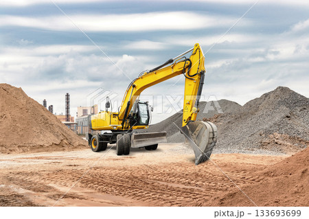 Yellow excavator operates on a construction site, moving large piles of sand and gravel under a cloudy sky, showcasing heavy machinery in action 133693699