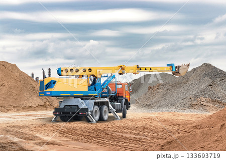 A yellow and blue crane truck lifts material at a construction site surrounded by large mounds of sand and gravel. Overcast skies add depth to the scene A yellow and blue crane truck lifts material at a construction site surrounded by large mounds of sand and gravel. Overcast skies add depth to the scene 133693719