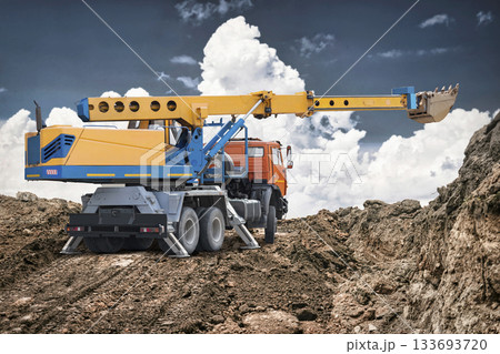 Construction workers operate a large crane and truck on a site with dirt piles and clouds in the background, indicating active work and progress 133693720