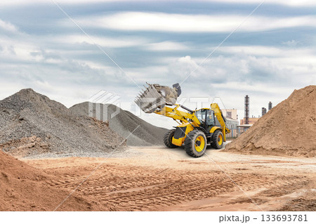 A yellow loader operates on a construction site, lifting sand and gravel with piles of materials nearby, against an industrial backdrop under gray clouds A yellow loader operates on a construction site, lifting sand and gravel with piles of materials nearby, against an industrial backdrop under gray clouds 133693781
