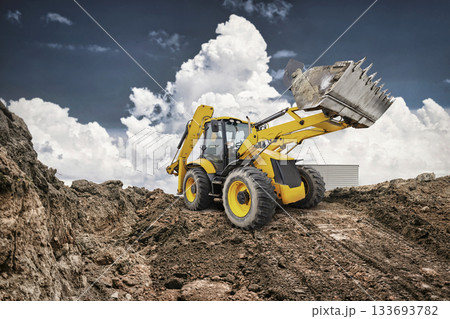 A yellow backhoe loader transports dirt at a construction site, highlighting the machinery's power against a backdrop of dramatic clouds A yellow backhoe loader transports dirt at a construction site, highlighting the machinery's power against a backdrop of dramatic clouds 133693782