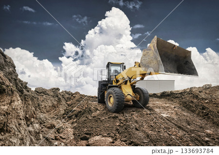 Heavy machinery is digging into the ground at a construction site. A loader is positioned against a backdrop of fluffy clouds and earth mounds 133693784