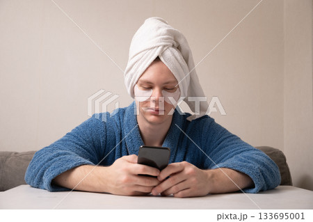 Young caucasian man wearing towel on his head and under-eye patches while using his smartphone during home skincare and relaxation routine. Mens Home Self-Care Routine 133695001