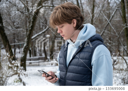 Side portrait of caucasian man checking phone while standing in winter park among bare trees. Outdoor Lifestyle 133695008