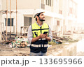 portrait of young engineer in vest with white helmet standing on construction site, smiling and holding smartphone for worker, internet, social media. 133695966