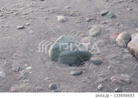 Smooth pebbles on a sandy shoreline, partially submerged in shallow water. The central pebble is distinctly larger, rounded, and grey, surrounded by smaller stones of various colors including muted re Smooth pebbles on a sandy shoreline, partially submerged in shallow water. The central pebble is distinctly larger, rounded, and grey, surrounded by smaller stones of various colors including muted re 133698683