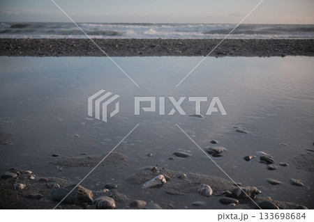 Smooth pebbles on a sandy shoreline, partially submerged in shallow water. The central pebble is distinctly larger, rounded, and grey, surrounded by smaller stones of various colors including muted re 133698684