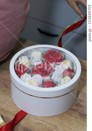 Female entrepreneur assembling marshmallow flowers in a hat box for a gift, tying it with colorful ribbons, marshmallow tulips and roses. Oblique Angle. Female entrepreneur assembling marshmallow flowers in a hat box for a gift, tying it with colorful ribbons, marshmallow tulips and roses. Oblique Angle. 133698705