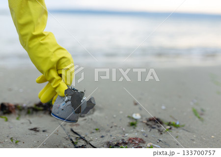 volunteer picking up a crushed metal can during beach cleanup, highlighting pollution, waste problem, ecology, coastal protection and environmental awareness. 133700757