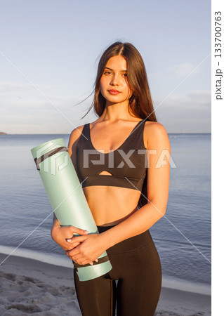 Woman holding yoga mat by the sea at sunset, fitness lifestyle, yoga training, outdoor wellness, healthy routine, seaside workout, calm evening yoga vibes. 133700763