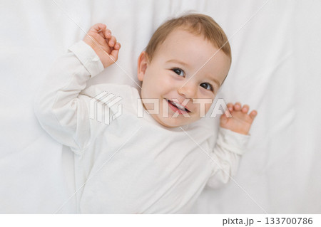 Cozy portrait of a happy baby lying on a soft white bed at home, smiling in natural light. A warm and cozy childhood moment filled with joy, innocence, comfort, and gentle family emotion. Cozy portrait of a happy baby lying on a soft white bed at home, smiling in natural light. A warm and cozy childhood moment filled with joy, innocence, comfort, and gentle family emotion. 133700786