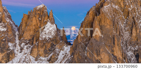 Panoramic view of Dolomite mountain peaks under a glowing full moon. Early winter landscape with snow-covered summits in soft dawn light. Captured in high resolution, ideal for large prints. 133700960