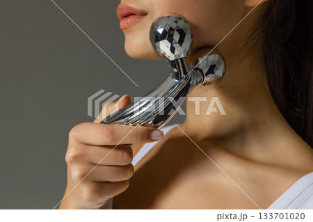 Close-up of a young woman massaging her neck with a metal 3D roller, demonstrating a tightening and relaxing skincare routine at home. 133701020
