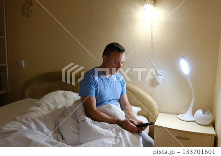 An adult male man sitting on a bed at night, smartphone screen, deep in thought. dim lighting and focused expression. Themes of insomnia, social media addiction, stress, late-night scrolling 133701601