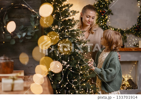 Caucasian mother and daughter decorating Christmas tree in cozy living room with warm lights Caucasian mother and daughter decorating Christmas tree in cozy living room with warm lights 133701914