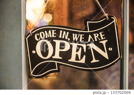 Closeup of a vintage open sign hanging on a shop window with warm bokeh lights in the background. 133702009