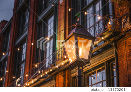 Warm street lantern glowing at dusk in Toronto's historic Distillery District, with festive string lights illuminating the old brick architecture. Warm street lantern glowing at dusk in Toronto's historic Distillery District, with festive string lights illuminating the old brick architecture. 133702014