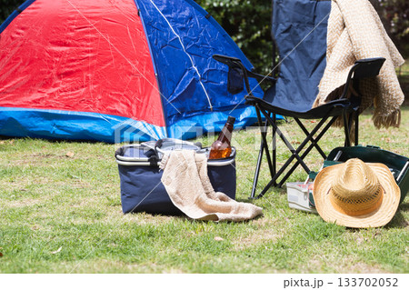 Outdoor scene featuring a red and blue camping tent pitched on green grass next to a folding chair, a cooler with a drink, and a straw hat, indicating a relaxing leisure activity 133702052
