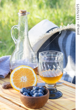 Outdoor picnic in the countryside with fresh blueberries in a bowl, sliced orange, glass of golden drink and vintage bottle on a wooden tray for a special moment Outdoor picnic in the countryside with fresh blueberries in a bowl, sliced orange, glass of golden drink and vintage bottle on a wooden tray for a special moment 133702075