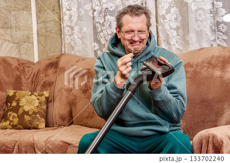 Scrubbing vacuum nozzle, middle aged man tries to clear debris from cleaning attachment, face twisted in visible disgust while doing chore. 133702240