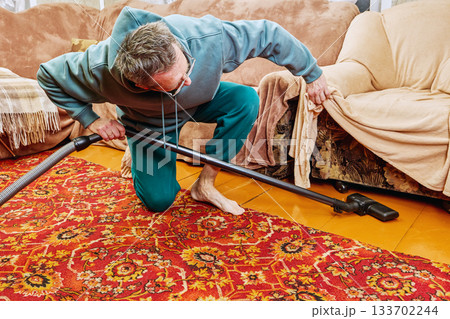 Man vacuuming floor in front of armchair covered with blanket, holding blanket with one hand. 133702244