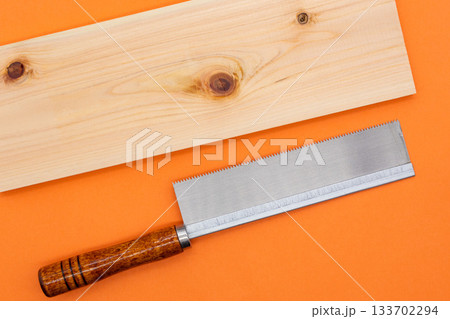 鮮やかなオレンジ背景に置かれた木材(ヒノキ材)と鋸(。日曜大工やDIY、リフォーム、木工の 鮮やかなオレンジ背景に置かれた木材(ヒノキ材)と鋸(。日曜大工やDIY、リフォーム、木工の 133702294