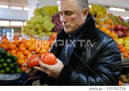 a man buys persimmons man with a beard shops for fresh fruit at an outdoor farmers' market. He is crouching and putting quince into a reusable mesh bag.  133702758