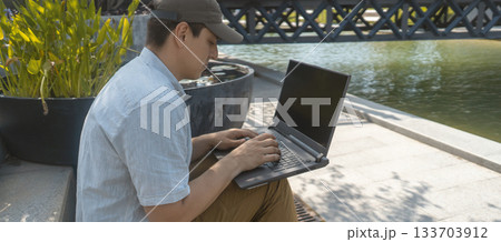 A man is sitting in a park and working on a laptop outdoors. A man is sitting in a park and working on a laptop outdoors. 133703912