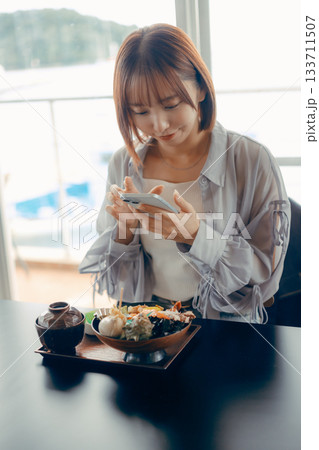 海鮮丼を前にスマホを操作する20代女性 海鮮丼を前にスマホを操作する20代女性 133711507